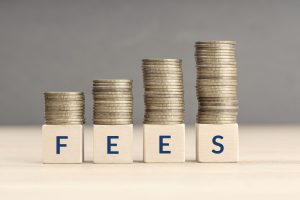 Fees increasing concept. Text word on wooden blocks with coins stacked in increasing stacks.